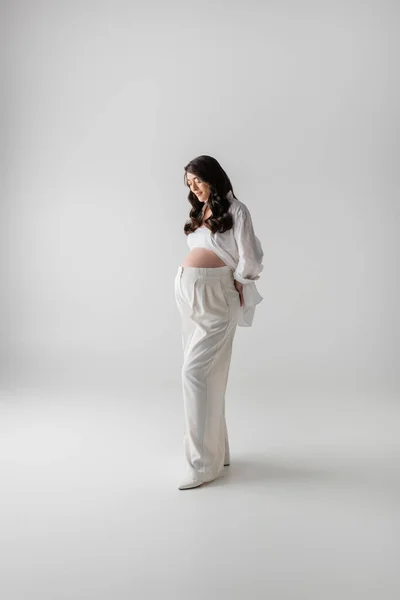 Piena lunghezza di bruna e attraente modello incinta in pantaloni eleganti bianchi e camicia che tiene la mano dietro la schiena mentre in piedi su sfondo grigio, concetto di maternità alla moda — Foto stock