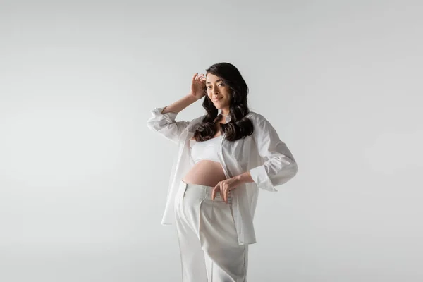 Femme enceinte à la mode avec des cheveux bruns ondulés posant en pantalon blanc et chemise tout en souriant à la caméra isolée sur fond gris, concept de style maternité — Photo de stock