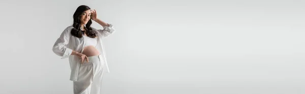 Hübsche und glückliche Schwangere in weißem Hemd, bauchfreiem Oberteil und Hose, die welliges brünettes Haar fixiert und isoliert auf grauem Hintergrund in die Kamera blickt, trendiges Umstandskonzept, Banner — Stockfoto