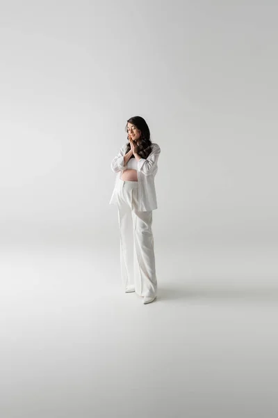Полная длина модного будущего мама в белых модных брюках и рубашке стоя с закрытыми глазами и улыбаясь на сером фоне, концепция материнства стиль, беременность — стоковое фото