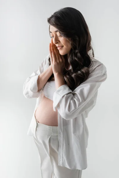 Charming and pleased pregnant woman with wavy brunette hair, wearing white crop top, shirt and pants, holding hands near face and smiling isolated on grey background, trendy maternity concept — Stock Photo