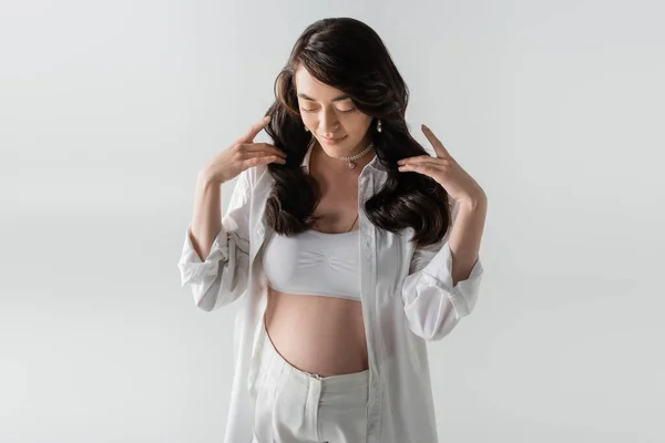 Fröhliche Schwangere in weißem Crop Top, stylischem Hemd und Hose, die welliges brünettes Haar berührt und isoliert auf grauem Hintergrund nach unten schaut, Umstandsmodekonzept — Stock Photo