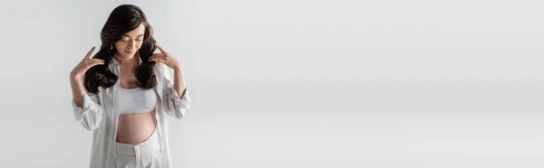 Stylische Schwangere in weißem, trendigem Hemd, bauchfreiem Oberteil und Hose, die welliges brünettes Haar berührt und isoliert auf grauem Hintergrund posiert, modisches Umstandskonzept, Banner — Stockfoto