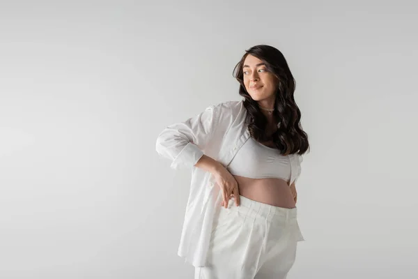 Studio shot of pleased future mother in white stylish shirt holding white pants, smiling and looking away isolated on grey background, maternity fashion concept — Stock Photo