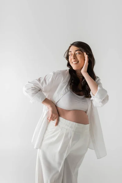 Stylish and charming pregnant woman in white crop top, stylish shirt and pants touching happy face and looking away isolated on grey background, maternity fashion concept — Stock Photo