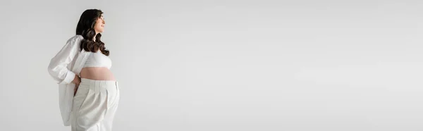 Vista laterale della modella incinta con capelli castani ondulati in posa bianca camicia elegante e pantaloni isolati su sfondo grigio, concetto di moda maternità, banner, moda maternità — Foto stock