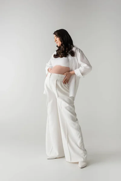 Longitud completa de atractiva y feliz futura madre en pantalones blancos de moda, top de la cosecha y camisa tocando vientre sobre fondo gris, maternidad de moda, embarazo - foto de stock