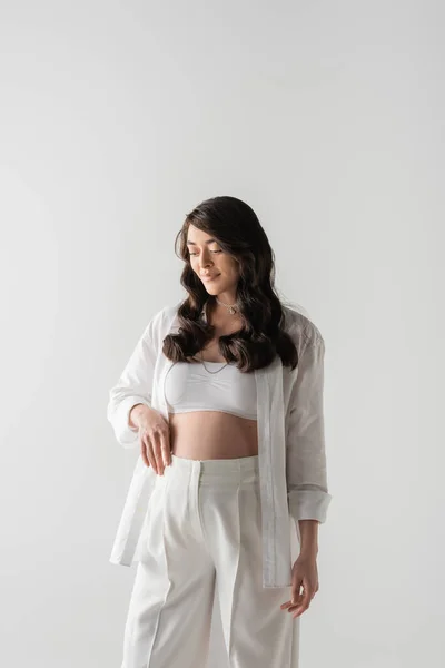 Задоволена вагітна жінка з хвилястим волоссям брюнетки, одягнена в білий верх, сорочка і штани, стоїть і посміхається ізольовано на сірому фоні, концепція моди материнства — стокове фото
