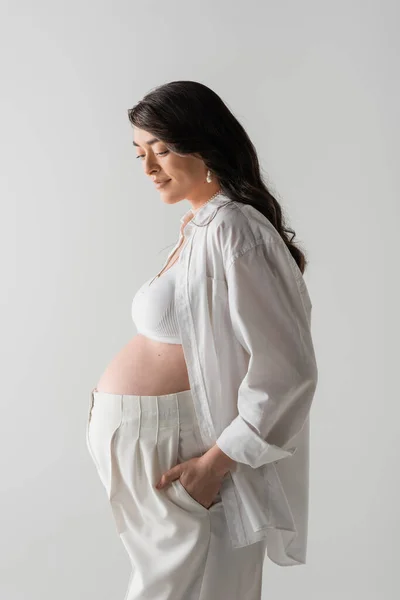 Side view of happy mom-to-be in white crop top, shirt and pants posing with hand in pocket, isolated on grey background, fashionable maternity concept, pregnant woman — Stock Photo