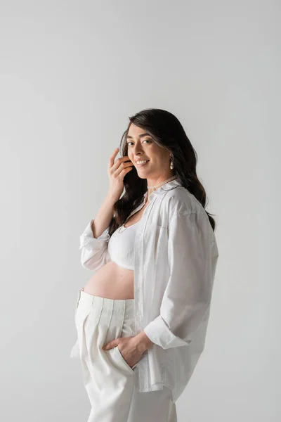 Mulher grávida encantadora com cabelo morena ondulado, em camisa branca, top de cultura e calças, sorrindo e olhando para a câmera isolada em fundo cinza, conceito de moda maternidade — Fotografia de Stock