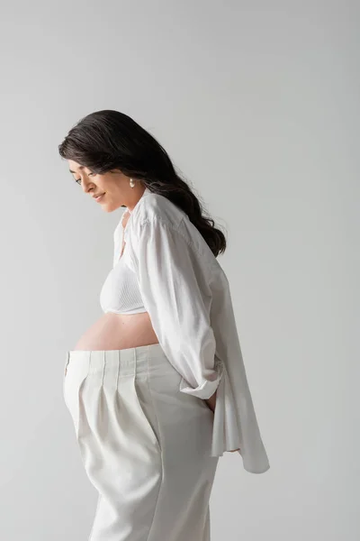 Side view of mom-to-be with wavy brunette hair, wearing white crop top, shirt and pants, posing with hand behind isolated on grey background, stylish pregnancy concept, pregnant woman — стоковое фото