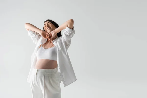 Brünette Schwangere in weißem Crop Top, Hemd und Hose, die Arme ausgestreckt und mit geschlossenen Augen auf grauem Hintergrund lächelnd, modisches Umstandskonzept — Stockfoto
