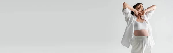 Allegro modello incinta in crop top bianco, camicia e pantaloni braccia tese con gli occhi chiusi isolati su sfondo grigio, concetto di moda maternità, banner — Foto stock