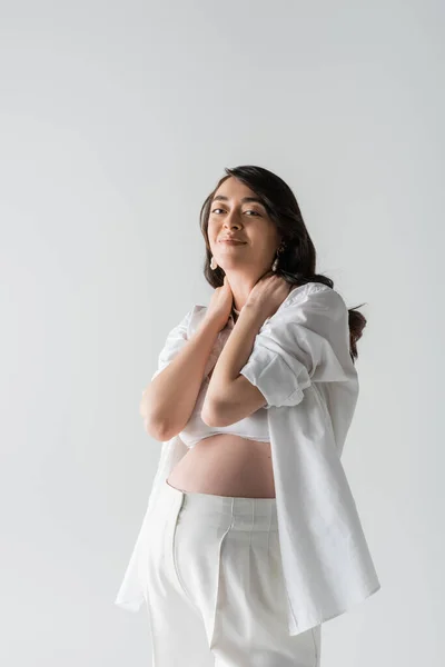 Joyful mother-to-be with wavy brunette hair touching neck and looking at camera while posing in white shirt and pants isolated on grey background, maternity style concept, pregnant woman — Stock Photo