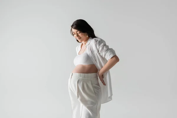 Happy future mother with wavy brunette hair, in stylish shirt, crop top and pants posing with hand behind back isolated on grey background, maternity fashion concept, pregnant woman — Stock Photo