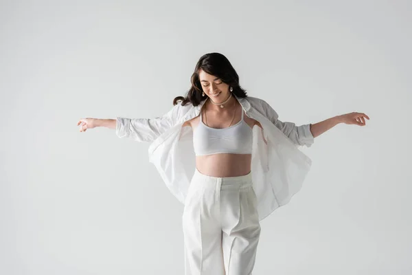 Cheerful and stylish pregnant woman in white crop top, pants and shirt standing with outstretched hands isolated on grey background, trendy maternity concept — Stock Photo