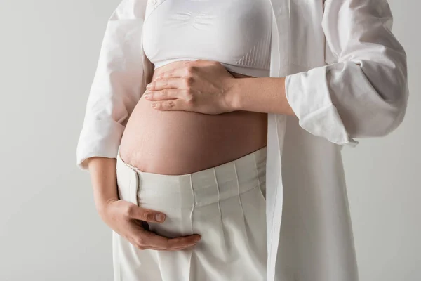 Partial view of future mother in white stylish shirt, crop top and pants tenderly embracing tummy isolated on grey background, maternity fashion concept, pregnant woman — Stock Photo