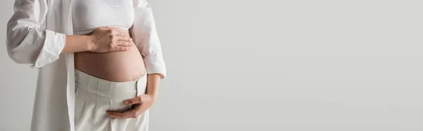 Частичный вид будущей матери в белом топе, рубашка и брюки, обнимающие живот, стоя изолированно на сером фоне, концепция материнства моды, баннер, беременная женщина — стоковое фото