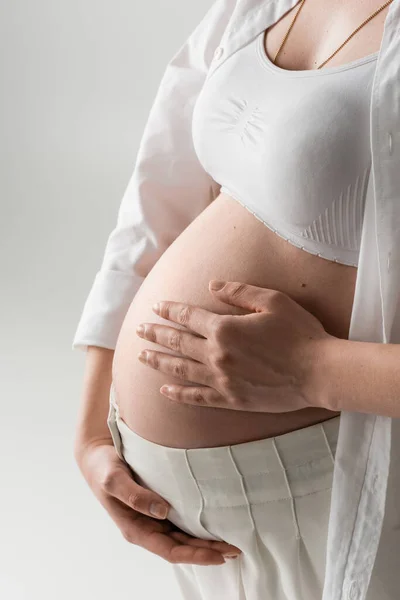 Cropped view of stylish expecting mother in white crop top, shirt and pants embracing tummy while standing isolated on grey background, maternity fashion concept, pregnant woman — Stock Photo