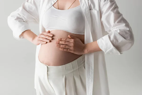 Частковий вид на модну очікувану матір у верхній частині врожаю, сорочка і штани позують руками на животику ізольовані на сірому фоні, концепція материнства, вагітна жінка — стокове фото