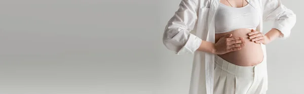 Vista recortada de la mujer embarazada de moda tocando el vientre mientras posa en la parte superior de la cosecha blanca, camisa y pantalones aislados sobre fondo gris, concepto de moda de maternidad, pancarta - foto de stock
