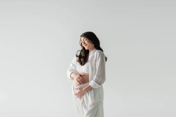 Appealing and stylish pregnant model in white crop top, shirt and pants embracing tummy and smiling with closed eyes isolated on grey background, maternity fashion concept — Stock Photo