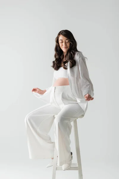 Charming mom-to-be with wavy brunette hair holding white shirt while posing on stool and smiling isolated on grey background, maternity fashion concept, pregnant woman — Stock Photo