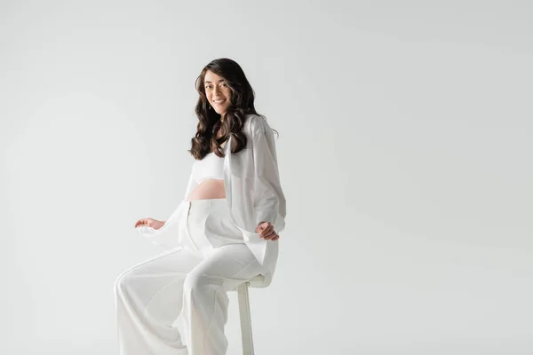 Cheerful pregnant woman in white fashionable clothes such as white shirt and pants sitting on stool and smiling at camera isolated on grey background, maternity fashion concept — Stock Photo