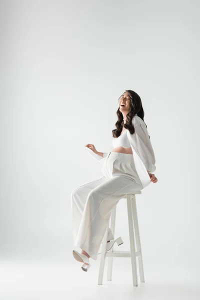 Full length of excited pregnant woman with wavy brunette hair sitting on stool in white trendy clothes and smiling with closed eyes on grey background, maternity fashion concept — Stock Photo