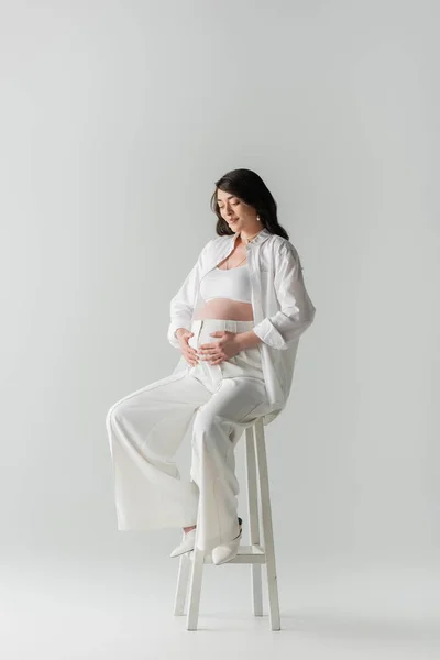 Full length of pleased future mother with wavy brunette hair smiling and touching belly while sitting on stool in white stylish clothes on grey background, maternity fashion concept, pregnant woman — Stock Photo