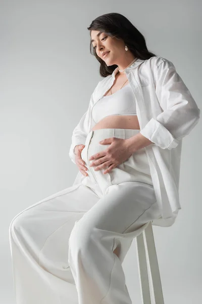 Smiling and appealing pregnant woman in white shirt, crop top and pants sitting on stool and embracing tummy isolated on grey background, maternity style concept, mother-to-be — Stock Photo