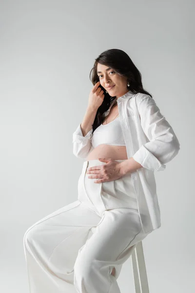 Encantadora mujer embarazada en ropa blanca de moda tocando el vientre y el pelo ondulado morena mientras sonríe a la cámara aislada sobre fondo gris, concepto de moda de maternidad - foto de stock