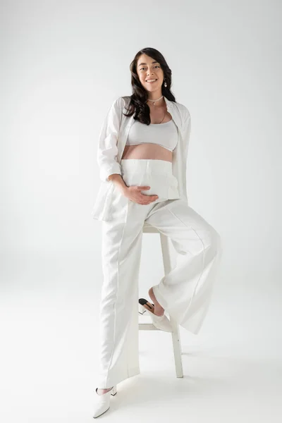 Повна довжина щасливої вагітної жінки в білому модному одязі, такому як верхній урожай, сорочка і штани, сидячи на табуреті і дивлячись на камеру на сірому фоні, модна концепція вагітності — стокове фото