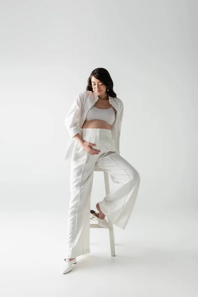 Full length of happy mom-to-be in white pants, crop top and shirt posing near stool and touching tummy on grey background, fashionable pregnancy concept, pregnant woman — Stock Photo