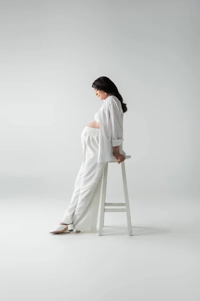 Side view of mother-to-be with wavy brunette hair posing near stool in white pants, crop top and shirt near stool on grey background, pregnancy fashion concept — Stock Photo