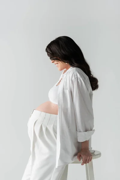Vista lateral de la mujer embarazada feliz con el pelo moreno ondulado posando en la parte superior de la cosecha blanca, camisa y pantalones aislados sobre fondo gris, concepto de maternidad de moda - foto de stock