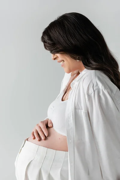 Side view of smiling pregnant woman in white crop top and shirt, face obscured with wavy brunette hair, touching belly isolated on grey background, maternity fashion concept — Stock Photo