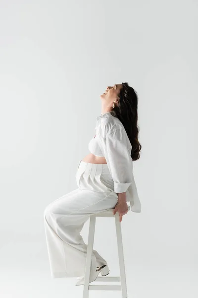 Side view of overjoyed pregnant woman with wavy brunette hair sitting on stool in white pants, shirt and crop top isolated on grey background, maternity style concept — Stock Photo