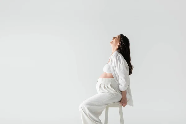Vue latérale de joyeuse femme enceinte avec des cheveux bruns ondulés assis sur des tabourets en chemise blanche, haut de culture et pantalon isolés sur fond gris, concept de maternité tendance — Photo de stock