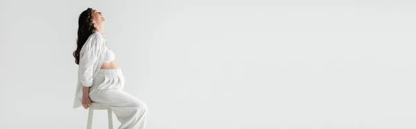 Vista lateral de feliz mom-to-be em calças elegantes, top de cultura e camisa sentada nas fezes e sorrindo isolado em fundo cinza, conceito de maternidade na moda, banner — Fotografia de Stock