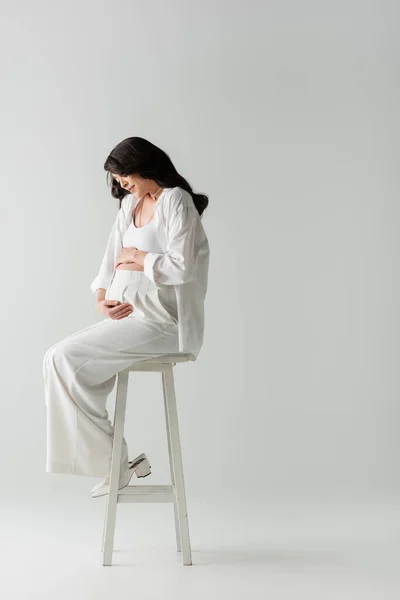 Pleine longueur de future mère élégante en chemise blanche et pantalon assis sur le tabouret et embrassant tendrement le ventre sur fond gris, concept de mode de maternité — Photo de stock
