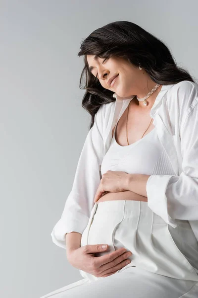 Overjoyed pregnant woman in white crop top, shirt and pants hugging belly and smiling with closed eyes isolated on grey background, maternity style concept — Stock Photo