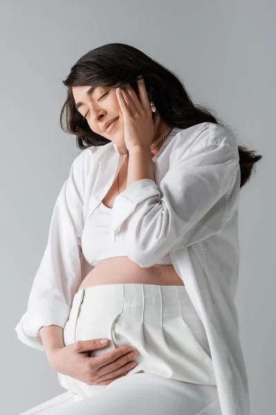 Morena mujer embarazada en top blanco, camisa y pantalones tocando vientre y cara feliz con los ojos cerrados aislados sobre fondo gris, concepto de estilo de maternidad - foto de stock