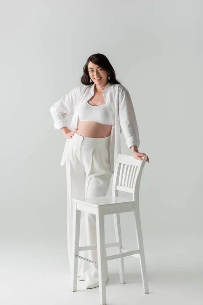 Full length of charming and cheerful mom-to-be in trendy pants, crop top and shirt posing with hand on hip near white chair on grey background, maternity fashion concept, pregnant woman — Stock Photo
