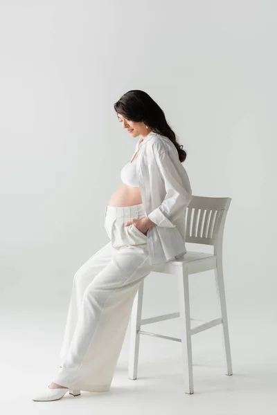 Повна довжина привабливої вагітної жінки у верхній частині врожаю і штани, що сидять на стільці з рукою в кишені штанів на сірому фоні, концепція материнства — стокове фото