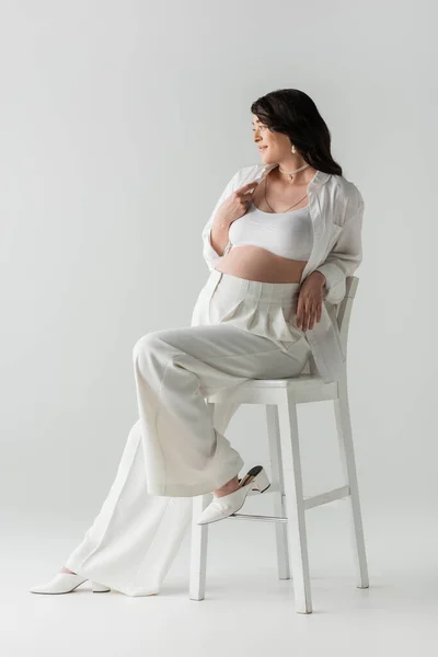 Полная длина модной беременной женщины в белом топе, рубашка и брюки сидя на стуле, улыбаясь и глядя в сторону на сером фоне, концепция моды материнства — стоковое фото