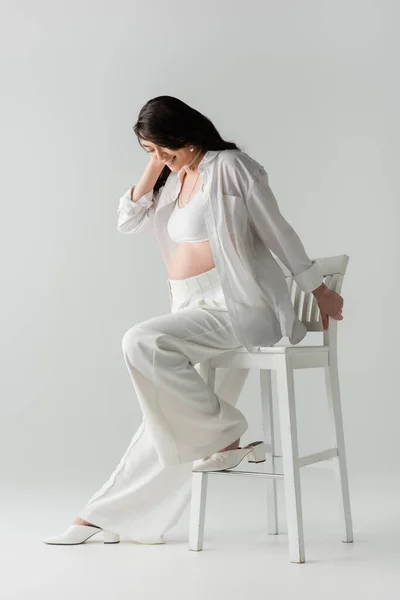 Longitud completa de complacido mamá-a-ser con el pelo ondulado morena sentado en la silla en camisa blanca, top de la cosecha y los pantalones en el fondo gris, concepto de maternidad de moda, mujer embarazada - foto de stock