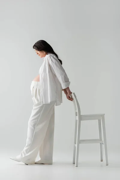 Vista lateral da mulher grávida morena e elegante em calças brancas, camisa e top da colheita posando com cadeira e sorrindo em fundo cinza, conceito de moda maternidade — Fotografia de Stock