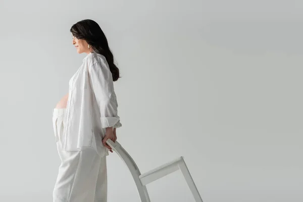 Vista lateral de la mujer embarazada de moda en camisa blanca y pantalones posando con silla mientras está de pie aislado sobre fondo gris, concepto de moda de maternidad - foto de stock
