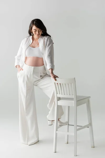 Повна довжина задоволеної майбутньої матері у верхній частині білого врожаю і сорочка, що стоїть з рукою в кишені штанів біля стільця на сірому фоні, трендова концепція вагітності — стокове фото